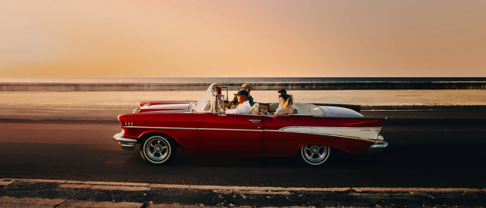 A classic convertible driving along a seafront road at sunset