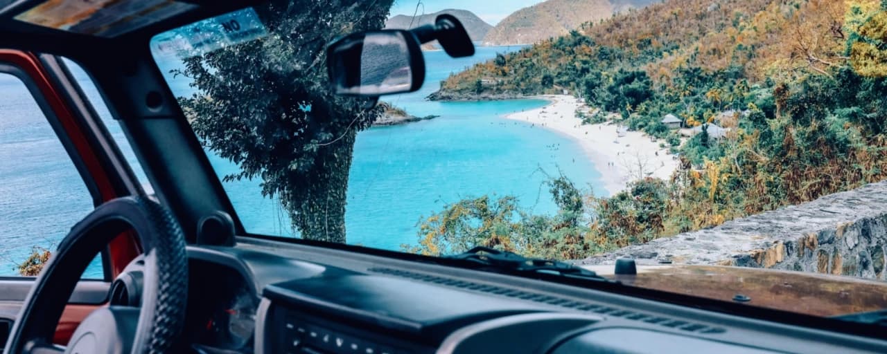 Driver's view over the wheel of a rental car looking out at a turquoise Caribbean bay
