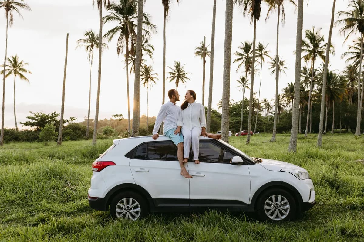 A couple sitting on the roof of a white SUV parked on grass among palm trees