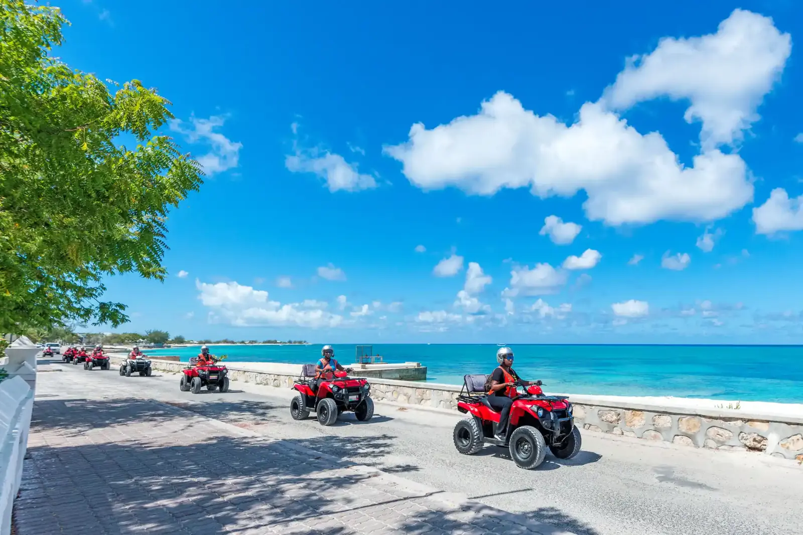 A guided group riding quad bikes along the oceanfront road beside the turquoise water in Cockburn Town, Grand Turk