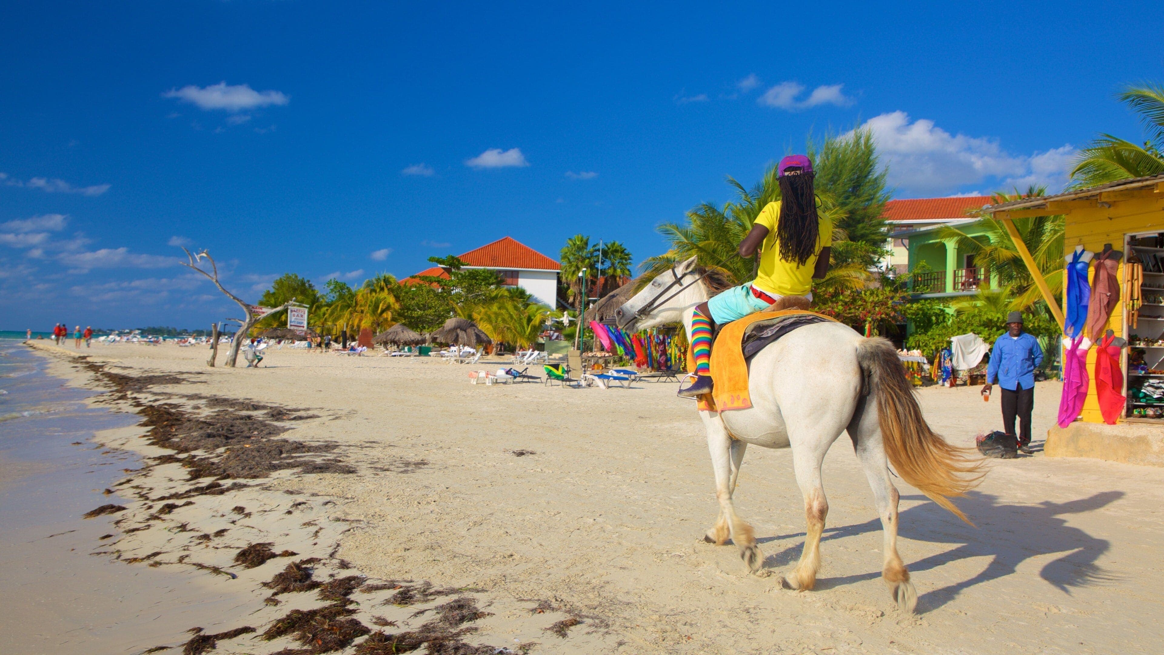 A rider on a white horse along the white sand of Seven Mile Beach with colourful beach shops in Negril, Jamaica