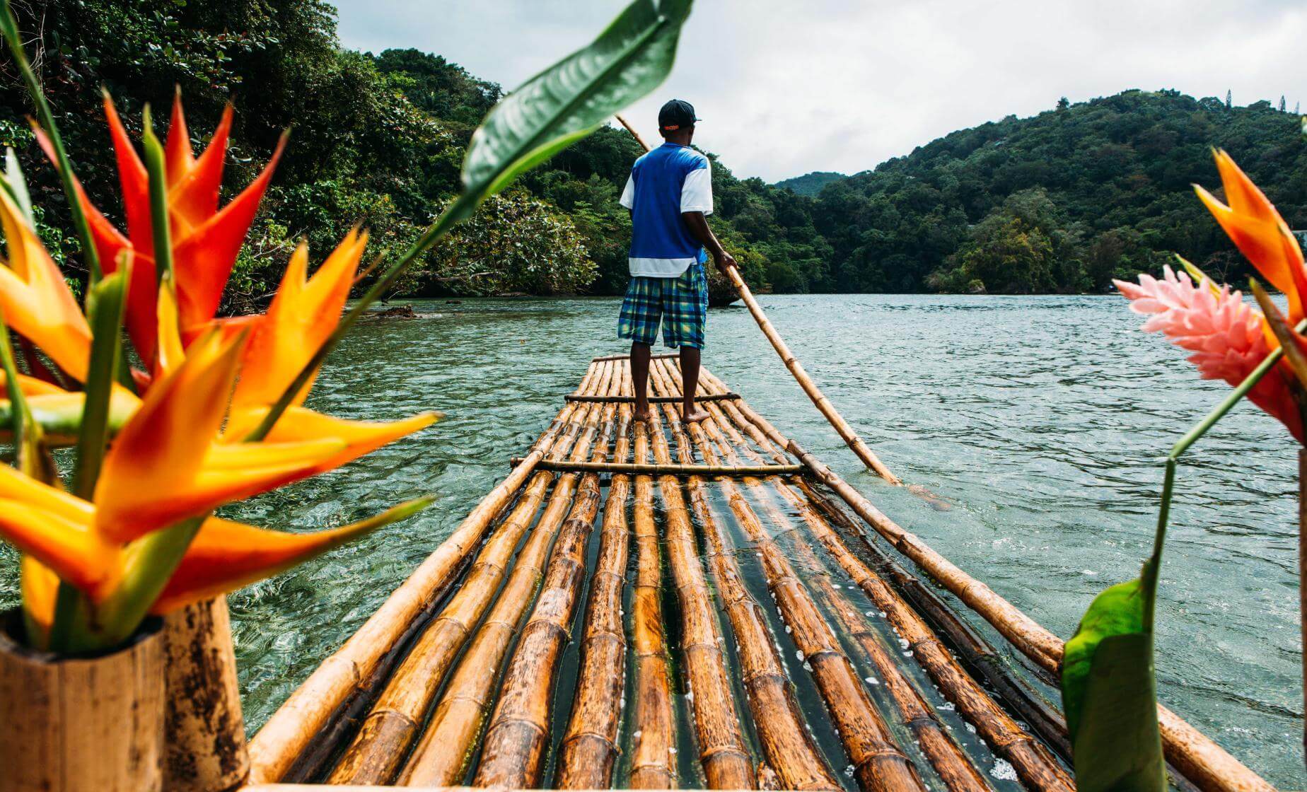A captain poling a bamboo raft down a calm jungle-lined river with tropical flowers in the foreground, Jamaica